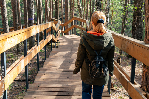 Young Woman Walking On Wooden Boardwalk Through Pine Forest With Backpack And Jacket