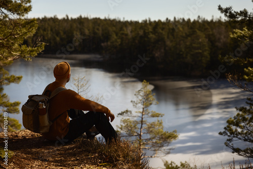 Canvas-taulu Hiker with a backpack and yellow beanie sitting on a cliff edge, enjoying the peaceful view of a frozen lake and dense evergreen forest during a sunny winter day in the quiet Scandinavian wilderness