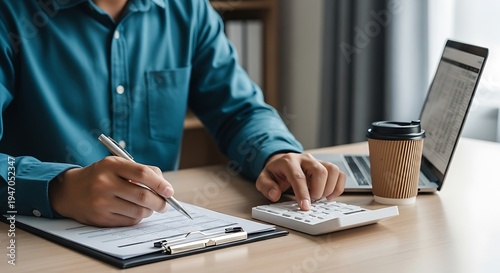 Man in a teal shirt working on paperwork with a calculator and laptop nearby daily