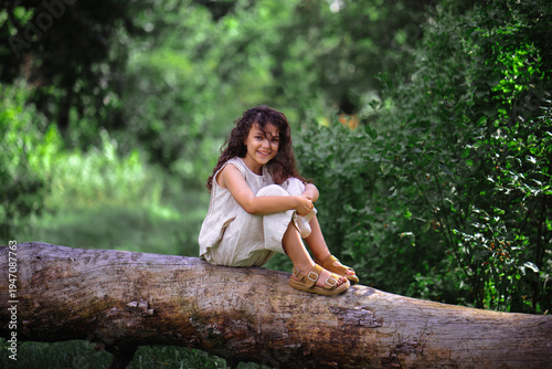 Happy little girl with curly hair smiling while sitting on a log in a sunny forest