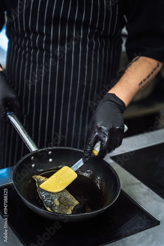 Professional chef in black gloves frying fresh fish fillet on a pan in restaurant kitchen