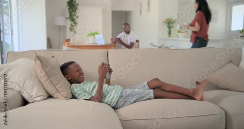 African American child on sofa in green shirt, watching tablet for fun while parents serving mugs