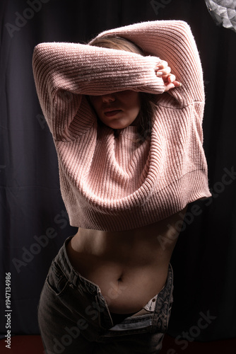 Female model with long dark hair poses in a pink ribbed sweater and high-waisted jeans against a dark backdrop, illuminated by soft overhead lighting