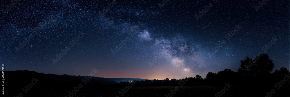 Fototapeta premium Milkyway panoramic night sky over horizon dark silhouetted trees and rolling fields wide celestial band and galactic core stretching across star filled sky