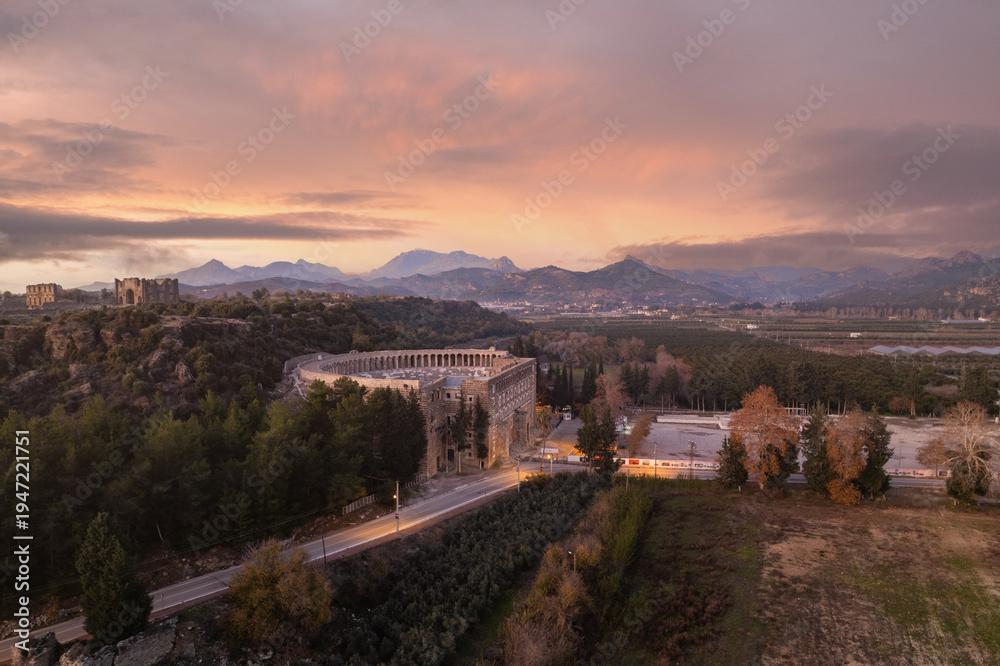Fototapeta premium Aerial view of the ancient Roman theater of Aspendos at sunset near Antalya, Türkiye, surrounded by Mediterranean landscape and dramatic colorful sky.