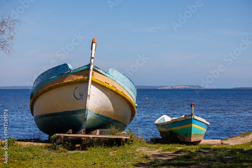 deux bateaux de pêche abandonnés sur un rivage, avec des traces de peinture écaillée et des inscriptions, placés sur des supports en bois à proximité de l'étang de Salses-Leucate, sous un ciel clair.