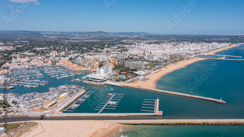 People visit Vilamoura marina in Portugal's Algarve region, enjoying boats and beach while the city skyline stands tall in the distance under clear blue sky Aerial
