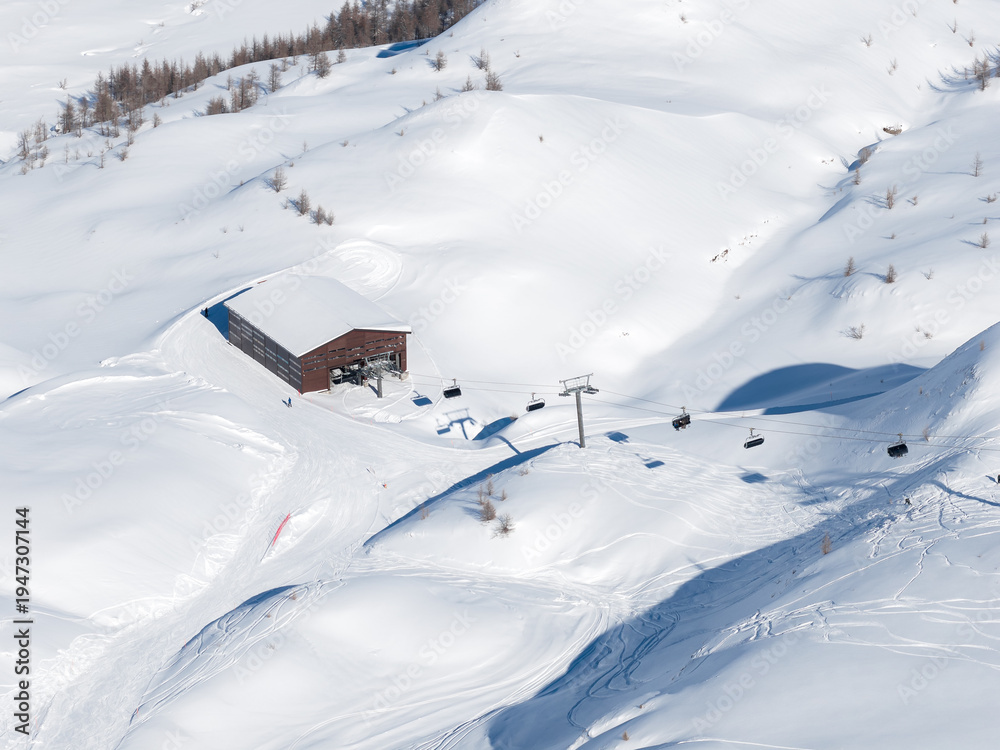 Fototapeta premium Aerial view of San Domenico ski resort in the Italian Alps shows a wooden lift station, a chairlift crossing powder, faint ski and snowboard tracks, and sparse larch trees.