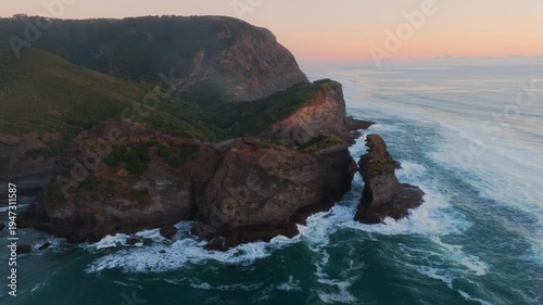 Aerial view of the rugged cliffs and sea stacks at Piha, Auckland, New Zealand, where the Tasman Sea meets the shore, creating a dramatic coastal landscape.