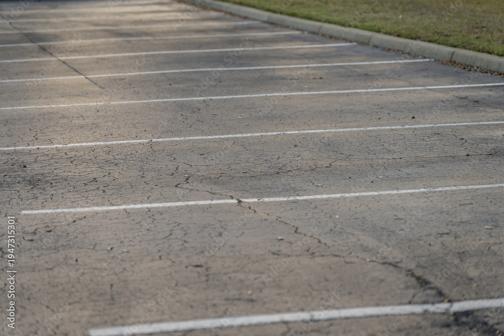 Fototapeta premium Empty parking spaces with white painted bay lines across cracked asphalt, bordered by a curb and grass strip in a quiet outdoor daylight scene