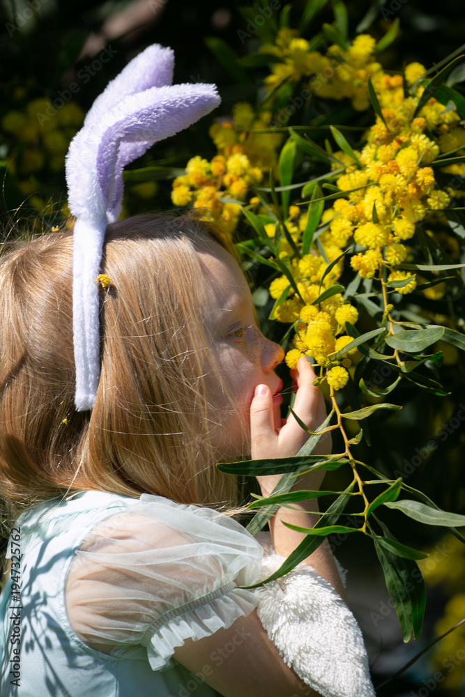 Naklejka premium A young girl wearing bunny ears touches yellow mimosa flowers while holding a plush bunny during Easter