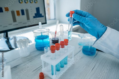 Close up of a gloved hand handling a test tube filled with blue chemical solution in a laboratory. Advanced technology and scientific research tools are present on the white worktable