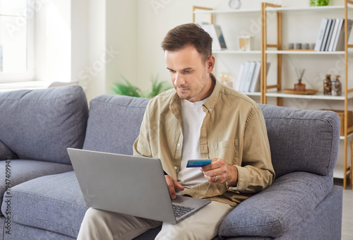 Man uses laptop, credit card for online payment. He verifies banking details and enters checkout data on the sofa at home, focusing on ecommerce shopping and finance. Internet banking concept.