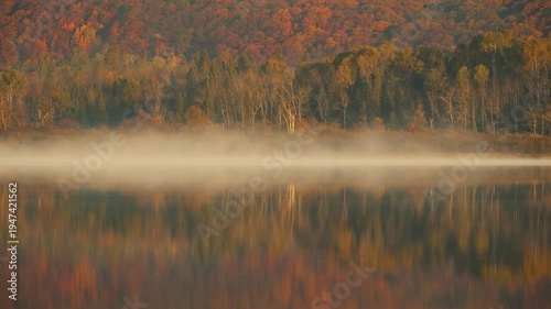 Morning fog rising over calm lake at sunrise with autumn forest reflection, ethereal mist moving across still water surface, cinematic fall nature timelapse