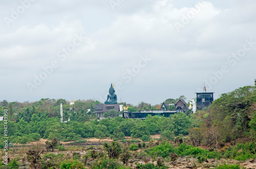 The large green buddha statue sits among lush green trees and buildings on a rocky hill under a cloudy sky