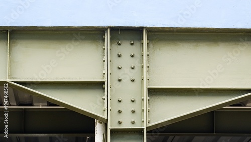 A detailed close-up of heavy-duty olive green steel beams and girders connected with industrial bolts and plates, forming a strong structural framework against a blue background.