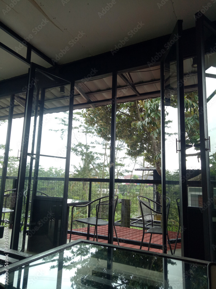Fototapeta premium Modern cafe interior with open glass doors to a balcony terrace, metal chairs and table, greenery and trees outside, soft natural light