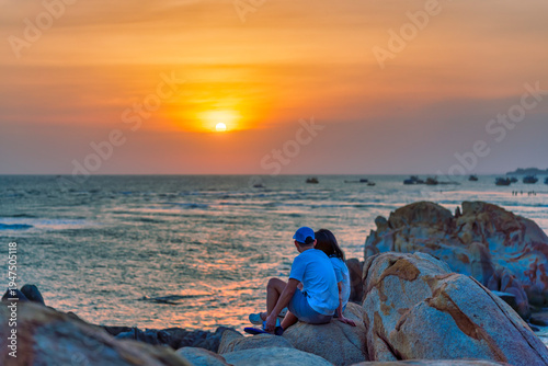 Wallpaper Mural Coastal sunset scene featuring a couple sitting together on large rocks by seaside. The sky glows with vibrant shades of orange, pink, and blue as the sun sets over the horizon Torontodigital.ca