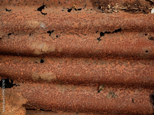 A close-up shot of heavily rusted and corroded corrugated iron, featuring deep orange oxidation and various holes caused by long-term weathering.