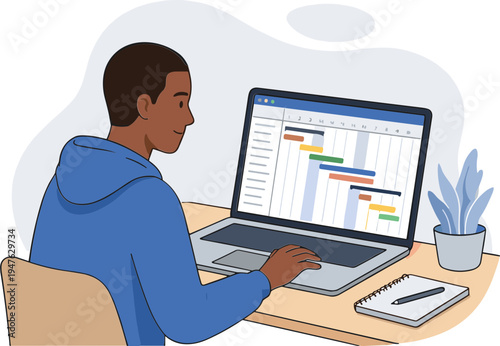 A man sits at a desk working on a laptop with a plant and notebook nearby.