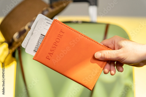 Person holds an orange passport holder and boarding pass near suitcase at the airport during daytime travel preparation