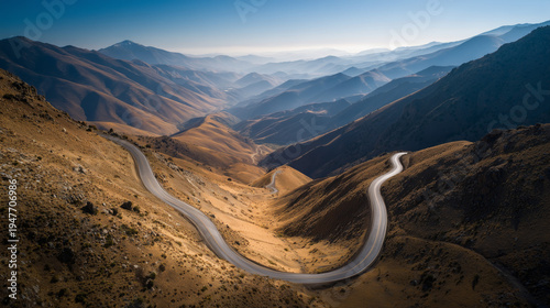 Winding mountain road through a vast, sunlit valley with layered hills and a clear blue sky.