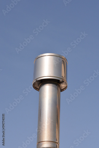 A shiny silver stainless steel chimney flue pipe with a cylindrical cap standing vertically against a clear blue sky background