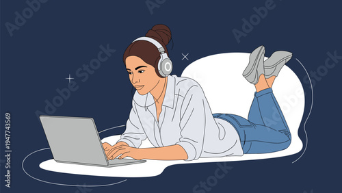 Young woman wearing headphones lies on her stomach while focused on her laptop screen for work or entertainment.