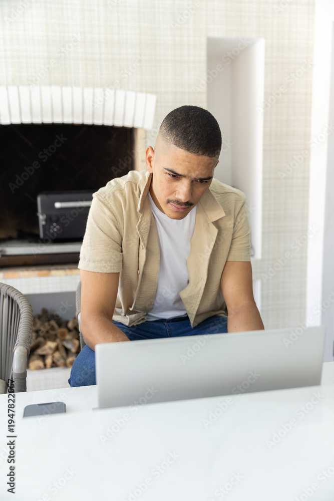 Fototapeta premium Man in his 30s sitting at home using laptop, smartphone left, wearing beige shirt, fireplace behind