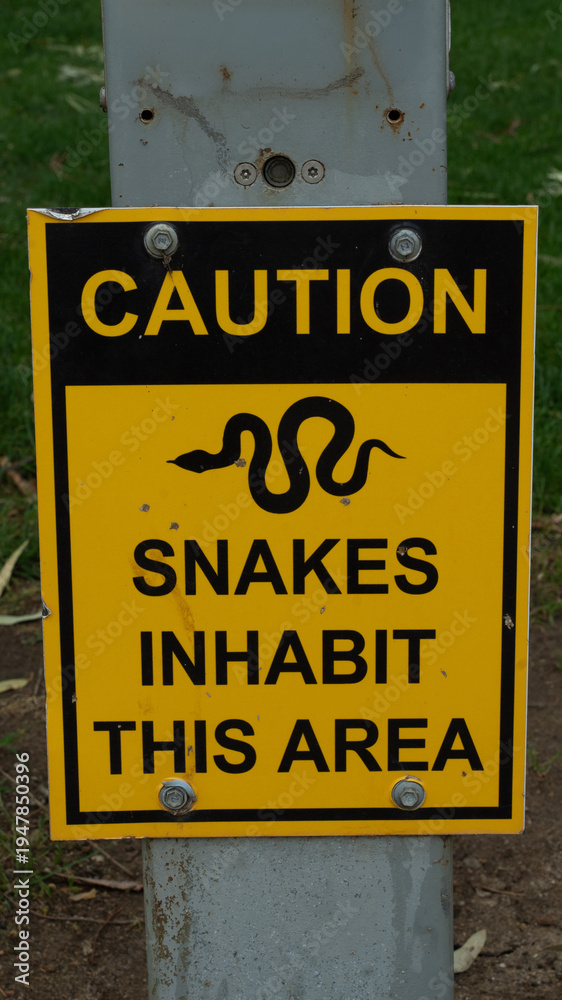 Fototapeta premium Yellow and black caution sign warning snakes inhabit this area mounted on metal post outdoors