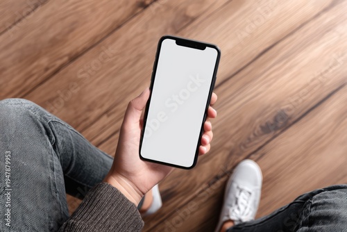 Person holding a modern smartphone with a blank white screen, viewed from above, against a wooden floor background.