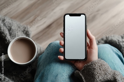 Overhead view of a person holding a smartphone with a blank screen, next to a cup of coffee.