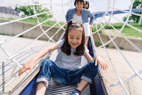 A young girl slides down a playground structure with her parents behind her, enjoying a fun day outdoors.