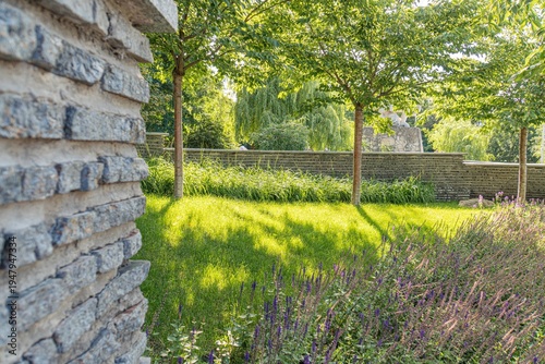 Old European city garden with stone wall, trimmed lawn, tall trees, purple flowers, natural light a