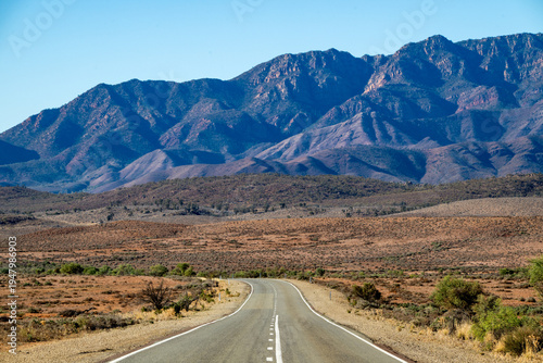 A long straight outback road leads towards a dramatic mountain range under a clear blue sky