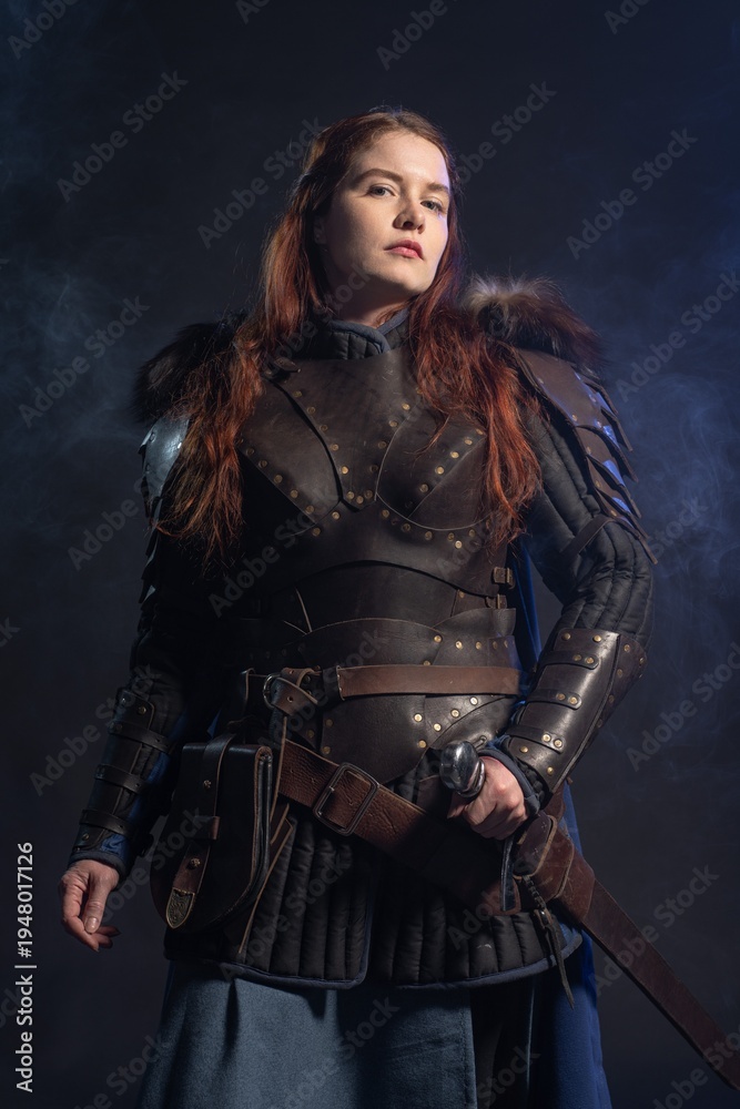 Fototapeta premium A fierce warrior woman with fiery hair stands ready for battle, clad in leather armor and a flowing blue skirt against a smoky backdrop.