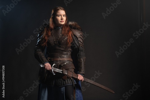 A fierce warrior woman stands poised with a sword against a dark backdrop, clad in armor and ready for battle.