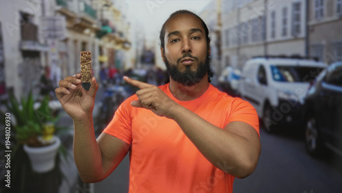 Canvas Print Man holding granola bar and points finger toward camera on urban street with parked vans and potted plants; healthy snack curiosity