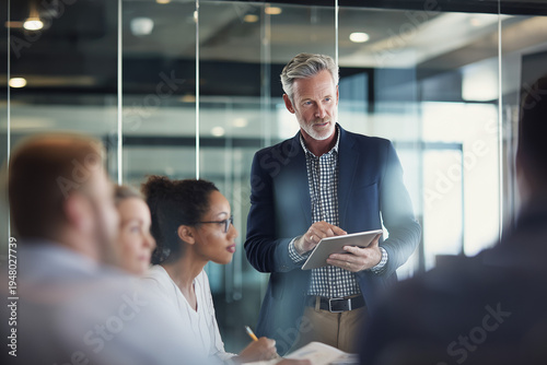 Team leader presenting ideas on digital tablet while coworkers listen and take notes in modern conference room.