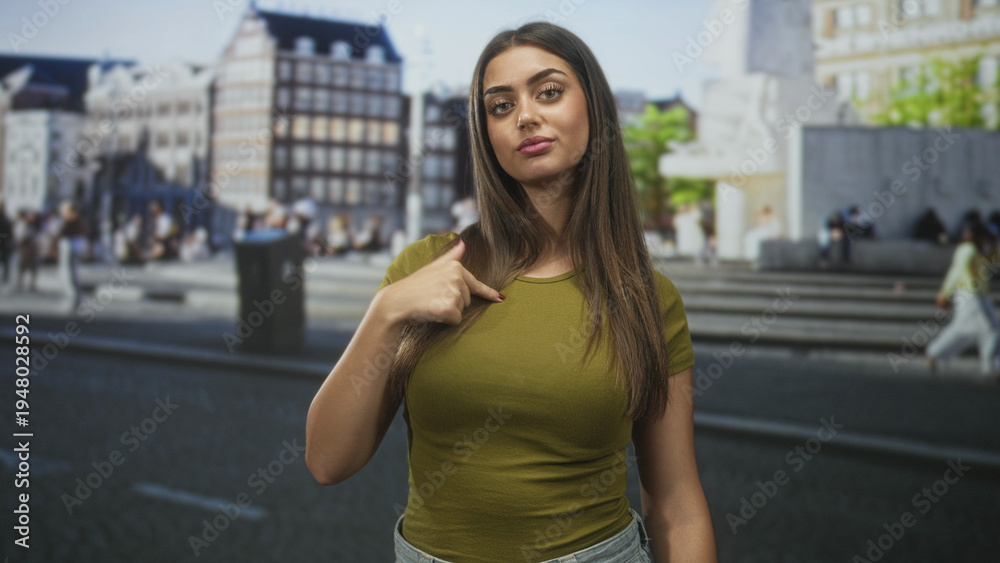 Fototapeta premium Woman points thumb to chest on a busy street in front of city buildings with a direct gaze at camera; confidence self identity.