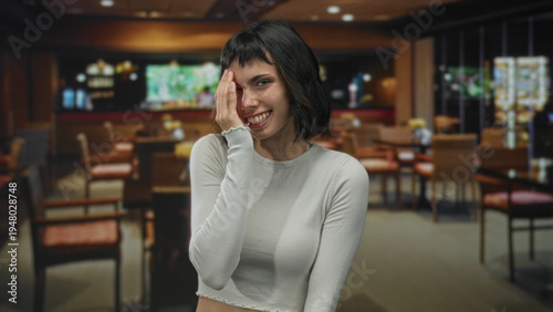 Young hispanic woman smiling broadly with hand covering face in a cozy restaurant building; embarrassment.