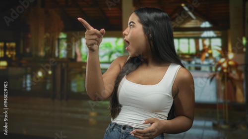 Tablou pe pânză Woman pointing finger with mouth open, wearing white tanktop and denim jeans, standing in a hotel lobby building near reception desk and plants, expressive gesture; surprise