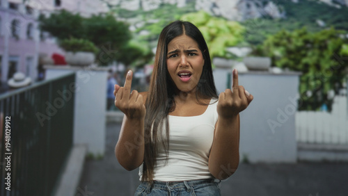 Vászonkép Young woman in white tanktop and jeans shows middle fingers and extended hands on a street near a railing; bold defiance