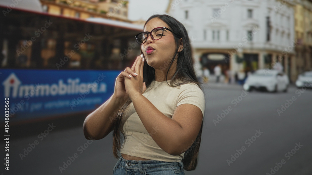 Fototapeta premium Young woman wearing glasses makes finger gun with hands aimed at camera on a busy city street with a red bus behind; playful confidence.