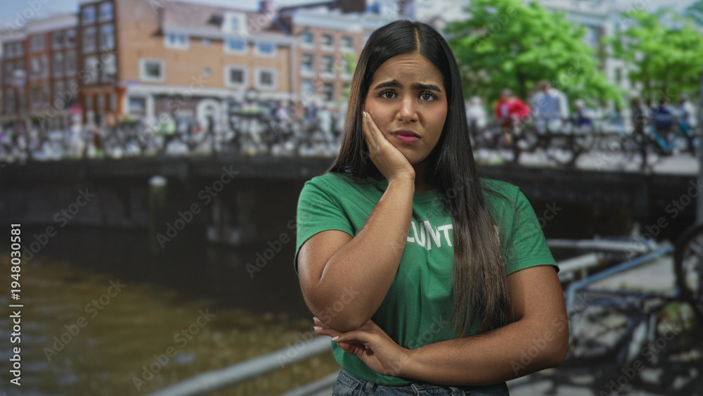 Fototapeta premium Young woman volunteer with hand on chin and crossed arms on a street by a canal and bicycles; concern reflection.