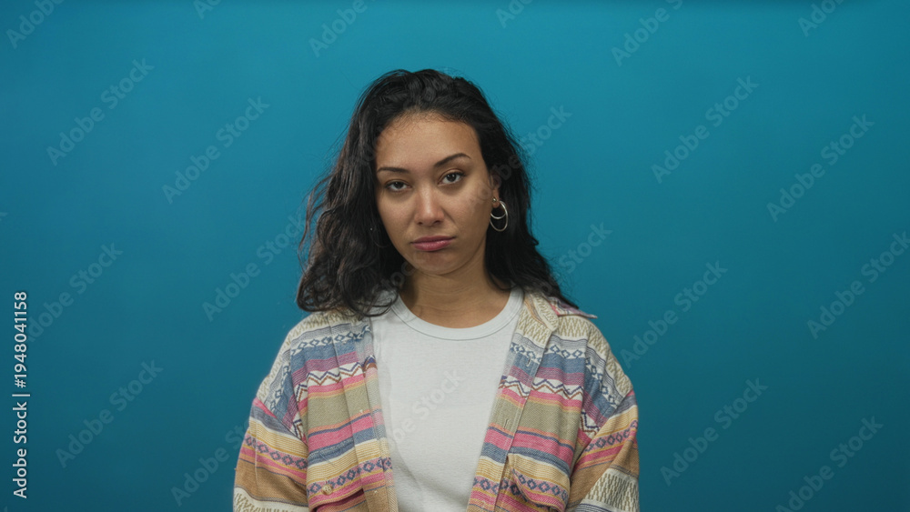 Fototapeta premium Young woman face glances up in studio against smooth teal backdrop wearing patterned cardigan; skepticism doubt contemplation.