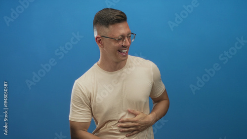 Konstfotografi Young man laughing with hand on chest and hand on stomach, wearing glasses and beige t shirt, against blue studio backdrop; joy warmth