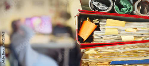 heap of file folders and blurred workplace with unrecognizable person in the background.Business concept,home office,freelance work,working at home,overworked, mental burnout,
