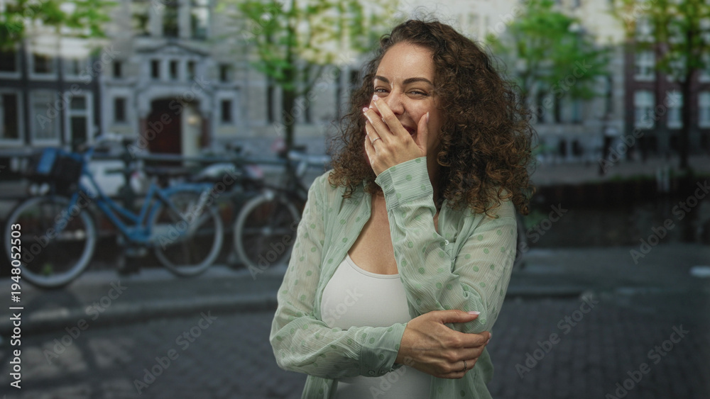 Fototapeta premium Woman laughing with hand covering mouth on amsterdam street by canal and bicycles; joy spontaneity now.