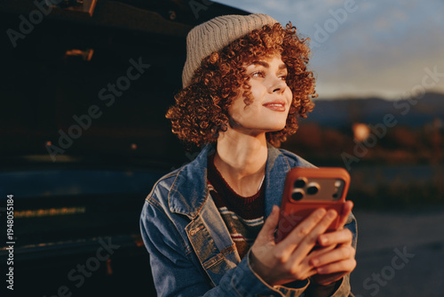 Curly woman wearing a beige hat and denim jacket holding smartphone while smiling outdoors during golden hour. Lifestyle concept with natural light and relaxed mood.